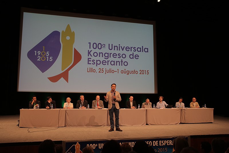 Dans un auditorium : Un homme debout fait un discours devant la salle, un panel est assis derrière lui.
